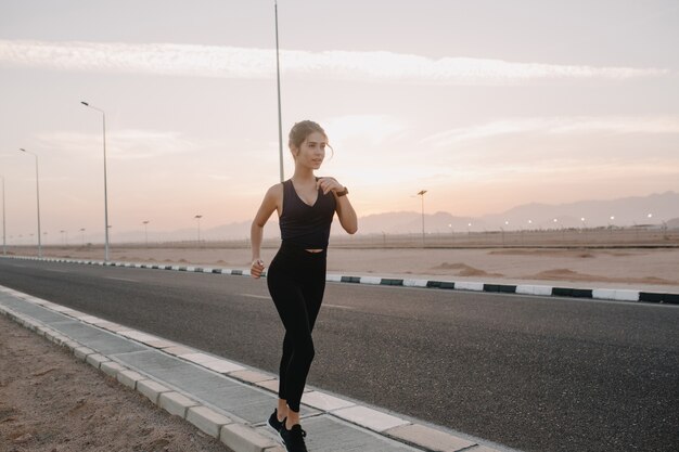 Treino no sol de manhã cedo na estrada do país tropical de mulher jovem e atraente no sportswear. Expressando positividade, emoções verdadeiras, estilo de vida saudável, treinamento, modelo forte