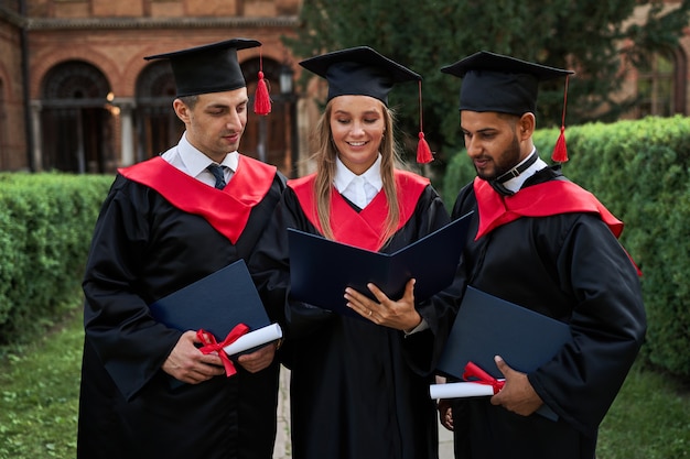 Três amigos de pós-graduação em vestes de formatura, olhando para seu diploma no campus.