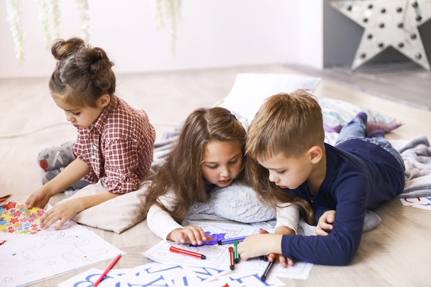 Três crianças focadas estão brincando no chão e desenhando livros para colorir