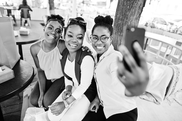 Três garotas afro-americanas casuais com sacolas de compras coloridas andando ao ar livre Mulheres negras elegantes comprando e fazendo selfie no telefone