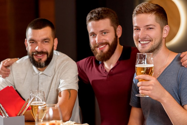 Três homens bonitos sorrindo, olhando para a câmera e segurando um copo de cerveja