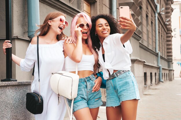 Três jovem e bela mulher hipster sorridente em roupas da moda de verãoMulheres multirraciais despreocupadas sexy posando no fundo da ruaModelos positivos se divertindo em óculos de sol tirando fotos de selfie