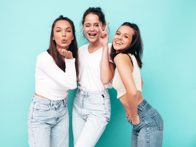 Foto grátis três jovem linda e sorridente morena hipster feminina em roupas da moda de verão mulheres despreocupadas sensuais posando perto da parede azul modelos positivos se divertindo alegre e feliz