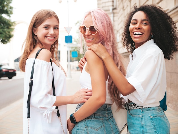 Três jovem linda mulher sorridente em roupas de verão da modaMulheres multirraciais despreocupadas sexy posando no fundo da ruaModelos positivos se divertindo em óculos de sol Alegre e feliz