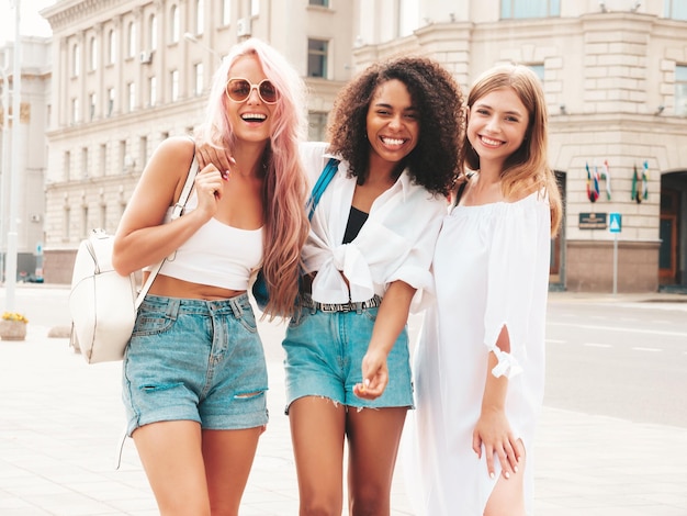 Três jovem linda sorridente hipster feminina em roupas da moda de verãoMulheres multirraciais despreocupadas sexy posando no fundo da ruaModelos positivos se divertindo em óculos de sol Alegre e feliz