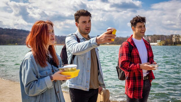 Três jovens amigos caminhando à beira do lago, bebendo e comendo em um parque