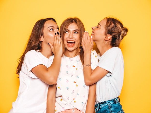 Três jovens bonitas garotas hipster sorridente em roupas da moda no verão. Mulheres despreocupadas "sexy" que levantam perto da parede amarela. Modelos positivos enlouquecendo e se divertindo. Compartilhe segredos, fofocas