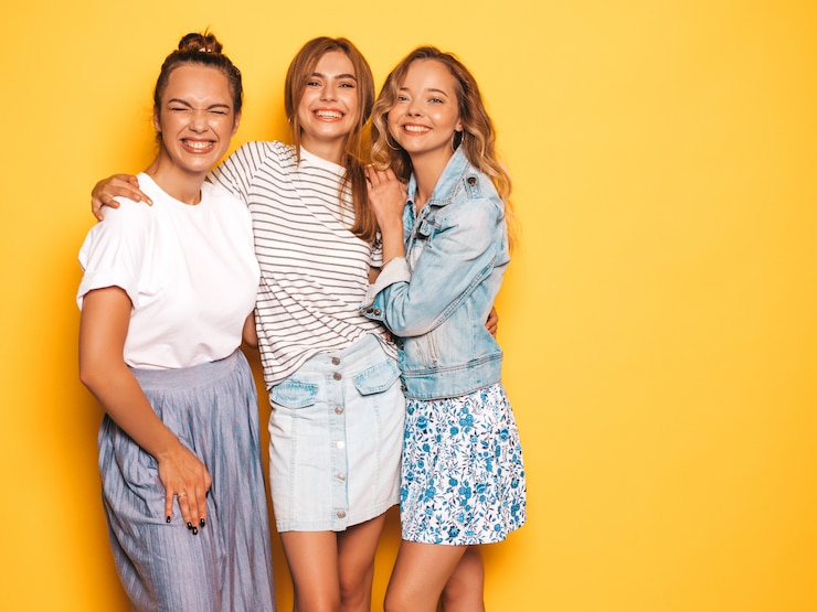 Foto grátis três jovens bonitas garotas hipster sorridente em roupas da moda no verão. mulheres despreocupadas "sexy" que levantam perto da parede amarela. modelos positivos se divertindo