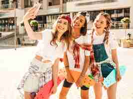 Foto grátis três jovens sorridentes meninas bonitas com skates centavo colorido. mulheres em roupas de hipster de verão posando no fundo da rua. modelos positivos tirando fotos de auto-retrato selfie em smartphone