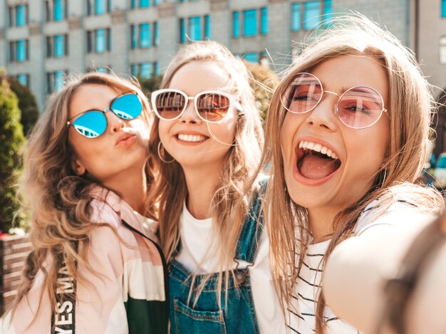 Foto grátis três jovens sorrindo hipster mulheres em roupas de verão. meninas tirando fotos de auto-retrato de selfie em smartphone. modelos posando na rua. feminino mostrando emoções de rosto positivo em óculos de sol