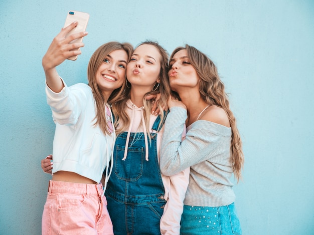 Três jovens sorrindo hipster mulheres em roupas de verão. meninas tirando fotos de auto-retrato de selfie em smartphone. modelos posando na rua perto da parede. feminino mostrando emoções de rosto positivo