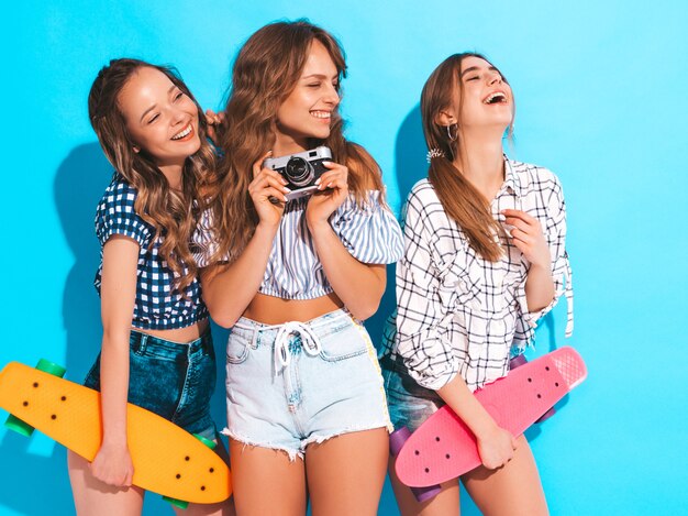 Três lindas meninas sorridentes elegantes com skates centavo coloridos. Mulheres no verão. Tirando fotos na câmera fotográfica retrô