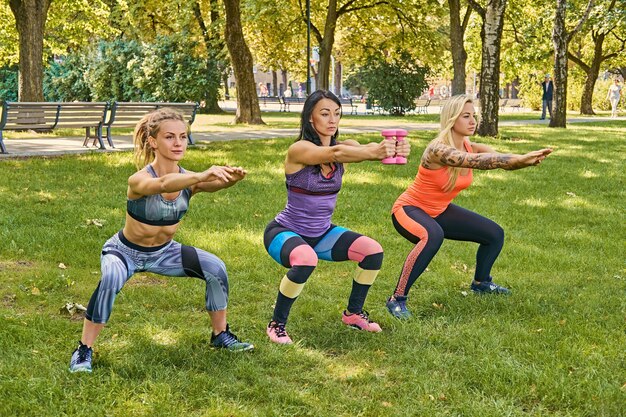 Três mulheres desportivas em roupas esportivas coloridas fazendo agachamentos em um parque.