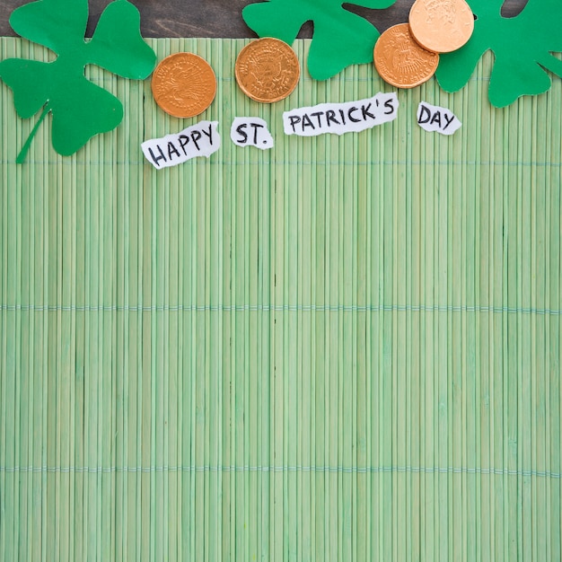 Trevos de papel perto de moedas e feliz título de st patricks day na esteira de bambu