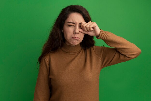 Triste, com os olhos fechados, bela jovem enxugando o olho com a mão isolada na parede verde