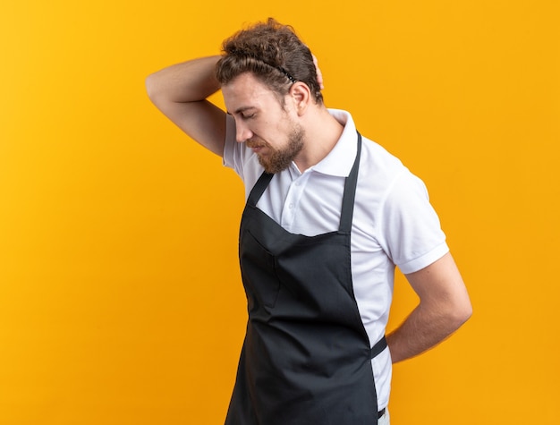 Triste com os olhos fechados, jovem barbeiro masculino vestindo uniforme, colocando a mão no pescoço isolado em fundo amarelo