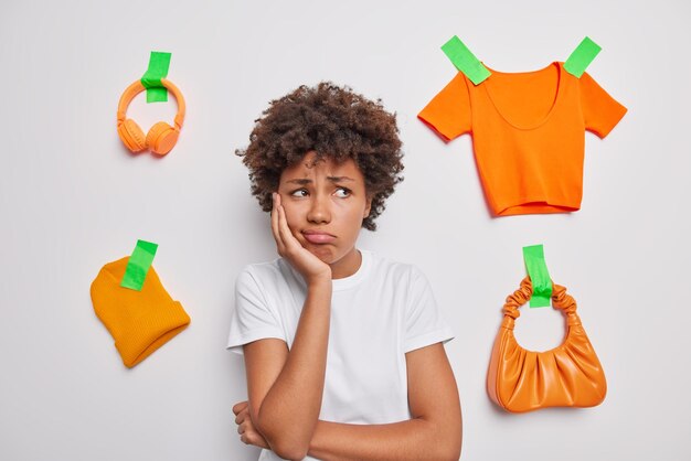 Triste descontente jovem de cabelos encaracolados desvia o olhar infeliz vestida com camiseta casual isolada sobre fundo branco com fones de ouvido laranja engessados chapéu camiseta e bolsa sendo profundo em pensamentos