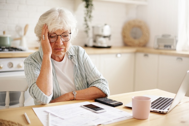 Triste e frustrada aposentada idosa com aparência deprimida, segurando a mão no rosto, calculando o orçamento familiar, sentada no balcão da cozinha com laptop, papéis, café, calculadora e telefone celular