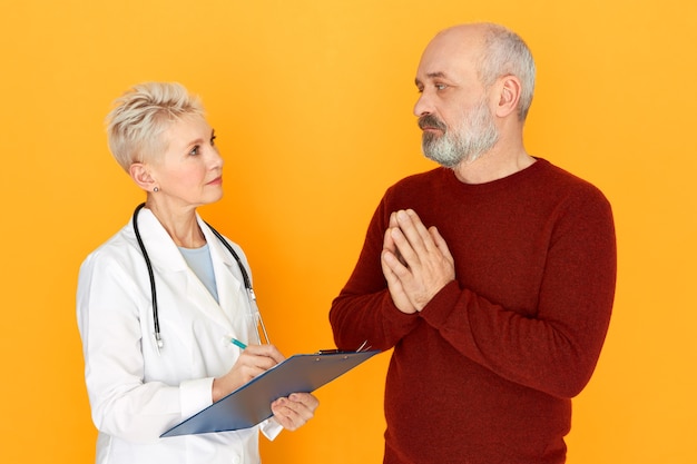 Triste e frustrado homem idoso com barba apertando as mãos pedindo a sua médica para curá-lo de uma doença respiratória durante o exame físico, falando sobre os sintomas.