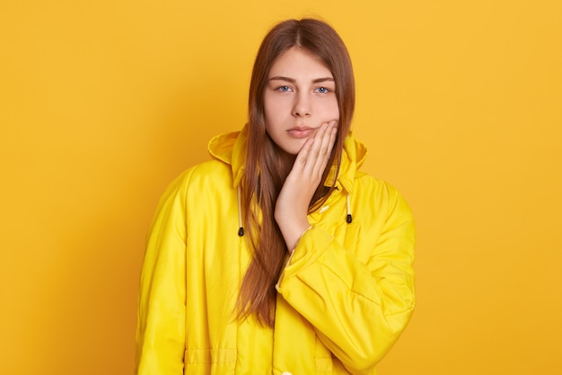 Triste fêmea vestindo jaqueta amarela tocando sua bochecha, sofrendo de dor de dente, tendo problemas médicos, de pé contra a parede amarela.