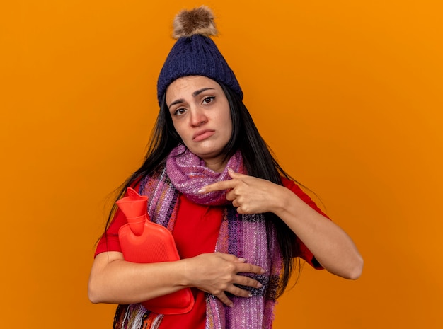 Foto grátis triste jovem caucasiana doente com chapéu de inverno e lenço segurando e apontando para uma bolsa de água quente isolada na parede laranja