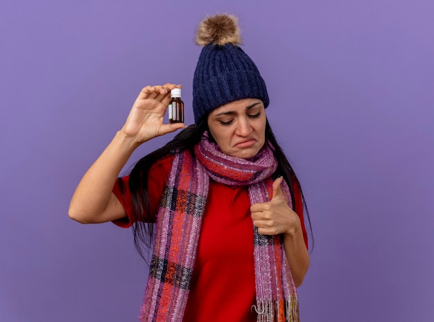 Triste jovem caucasiana doente com chapéu de inverno e lenço segurando medicamento em um vidro, olhando para baixo, mostrando o polegar isolado na parede roxa com espaço de cópia