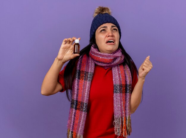 Triste jovem caucasiana doente usando chapéu de inverno e lenço segurando medicamento em um vidro, olhando para cima com o punho cerrado isolado no fundo roxo com espaço de cópia