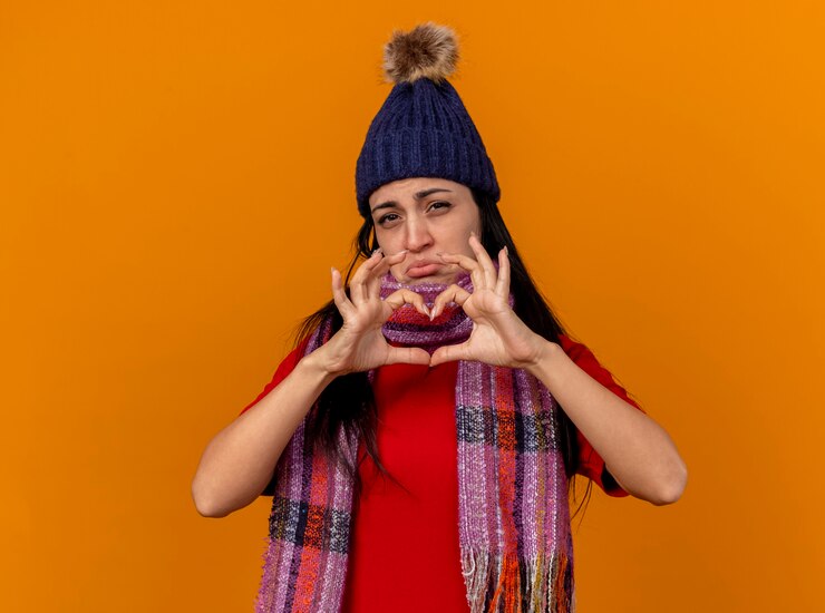 Foto grátis triste jovem caucasiana doente usando chapéu e lenço de inverno, fazendo um sinal de coração, olhando para a câmera isolada em um fundo laranja com espaço de cópia