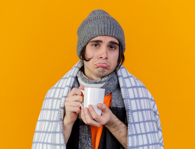 Foto grátis triste jovem doente usando chapéu de inverno com lenço embrulhado em xadrez segurando uma xícara de chá isolada em laranja