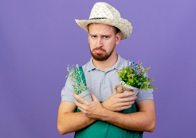 Triste jovem e bonito jardineiro eslavo de uniforme e chapéu olhando, de mãos cruzadas, segurando vasos de flores isolados