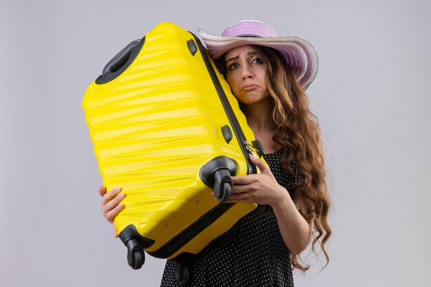 Triste jovem e linda garota viajante em um vestido de bolinhas com chapéu de verão, segurando uma mala, olhando para a câmera com uma expressão triste no rosto, de pé sobre um fundo branco