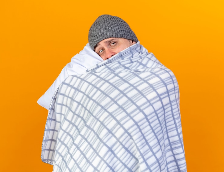 Foto grátis triste jovem loira doente com um chapéu de inverno embrulhado em uma manta segura e coloca a cabeça no travesseiro isolada na parede laranja