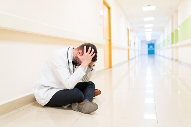 Triste médico adulto médio com a cabeça nas mãos, sentado no chão contra a parede no corredor do hospital