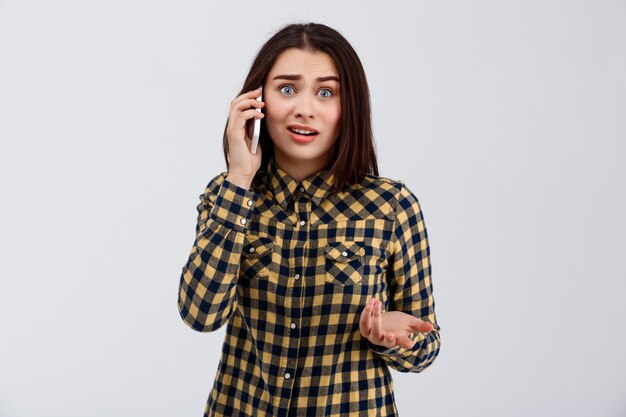 Triste menina bonita jovem vestida com camisa xadrez, falando no telefone, sobre parede branca.