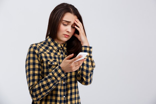 Triste menina bonita jovem vestida com camisa xadrez, segurando o telefone, olhando para a tela sobre parede branca.