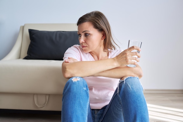 Triste mulher de meia idade sentada em casa no chão com um copo de água. problemas de saúde de mulheres mais velhas, saúde mental, dor de enxaqueca, sintoma de menopausa, período de depressão após coronavírus covid-19