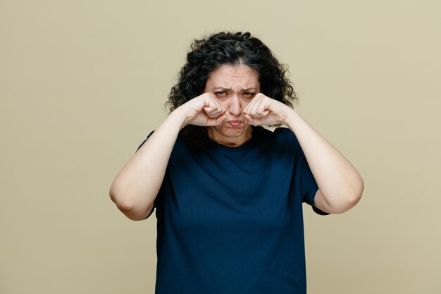 Triste mulher de meia idade vestindo camiseta olhando para a câmera fazendo gesto de choro isolado em fundo verde-oliva