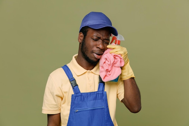 Triste segurando agente de limpeza com pano jovem limpador afro-americano de uniforme com luvas isoladas em fundo verde