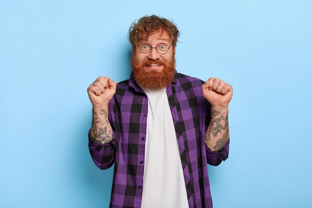 Foto grátis triunfando alegre homem com cabelo vermelho, levanta os punhos cerrados, comemora vitória ou triunfo, usa óculos ópticos