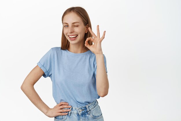 Tudo bem, sem problemas. Mulher loira sorridente asseguro-lhe, mostre sinal de OK OK e piscando satisfeito, elogie o excelente trabalho, bom trabalho, de pé na camiseta sobre fundo branco.