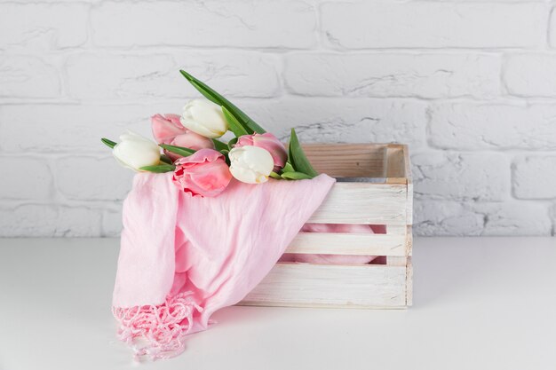 Tulipas e lenço rosa dentro da caixa de madeira na mesa contra a parede de tijolos brancos