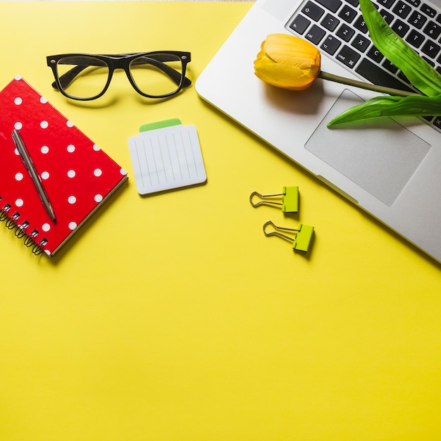 Tulipas no laptop com diário; caneta; óculos e clipes de papel sobre fundo amarelo