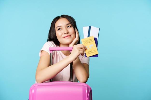 Turista asiática sonhadora, sorrindo e pensando, segurando um certificado internacional de vacinação covid ...