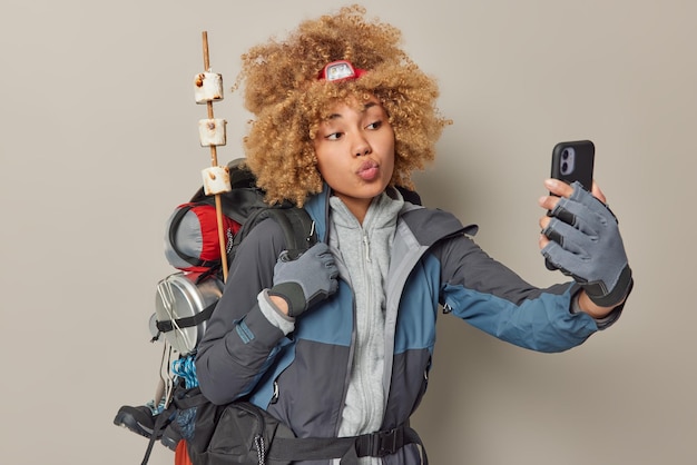 Turista ativo tira foto selfie mantém os lábios dobrados vestido com roupas esportivas carrega mochila pesada leva estilo de vida ativo tem expedição de viagem isolada sobre parede cinza Alpinista feminina retorna do piquenique