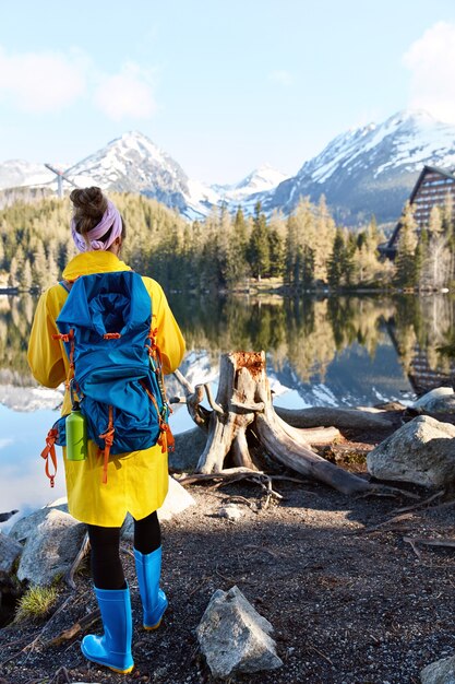 Turista com mochila perto do rio de montanha, aprecia a natureza selvagem com bela vista, usa anoraque longo amarelo