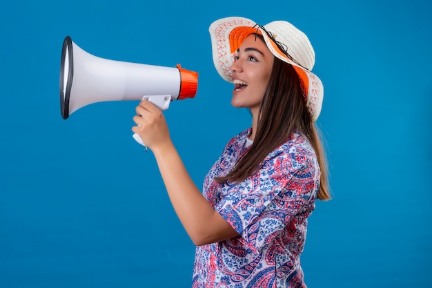 Foto grátis turista de jovem mulher bonita no chapéu do verão falando ao megafone com cara feliz sobre parede azul isolada