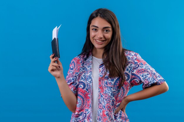 Turista jovem e linda segurando passaporte com ingressos, olhando para a câmera, sorrindo alegremente, positiva e feliz pronta para o feriado em pé sobre um fundo azul isolado