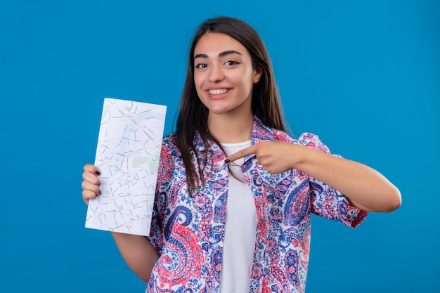 Turista jovem e linda segurando um mapa, olhando para a câmera com uma cara feliz apontando para ela com o dedo indicador e sorrindo alegremente em pé sobre um fundo azul isolado