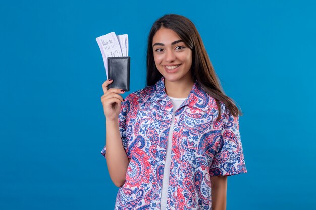 Turista jovem segurando passaporte com ingressos, olhando para a câmera, sorrindo alegremente, pronta para o feriado em pé sobre fundo azul isolado