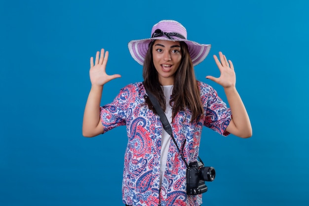turista mulher com chapéu e câmera fotográfica levantando as mãos em sinal de rendição parecendo surpresa em pé no isolado azul